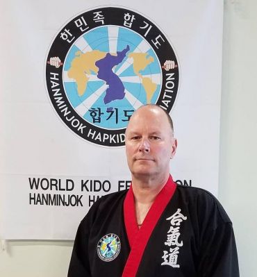 Claude T. Sullivan in a black uniform with a red collar, standing in front of the Hanminjok Hapkido Association banner.